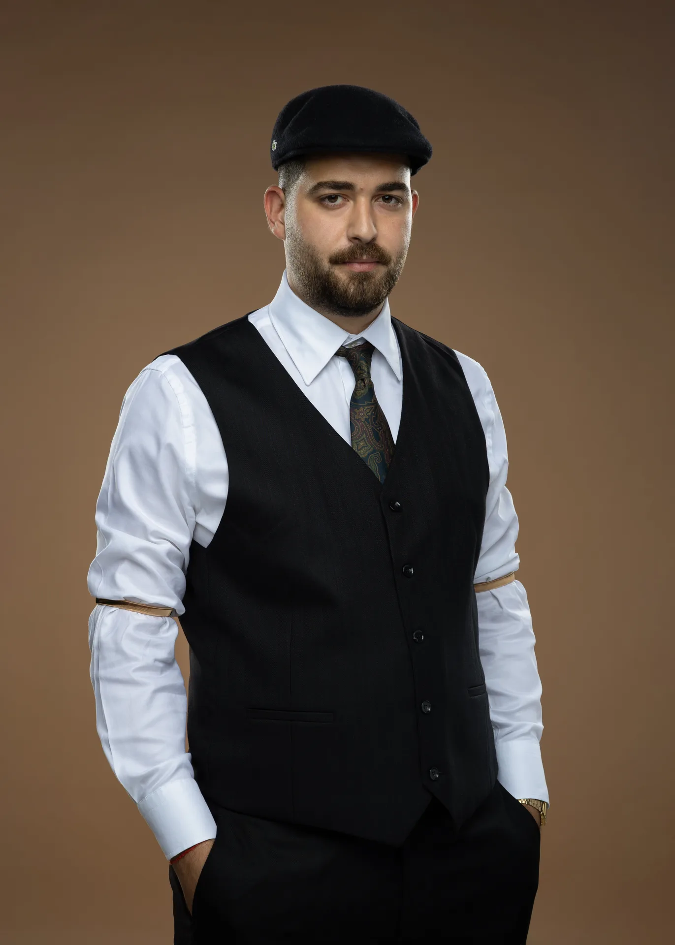 Portrait of a man in a white shirt, black waistcoat, and patterned tie wearing a black beret against a brown background, looking at the camera.