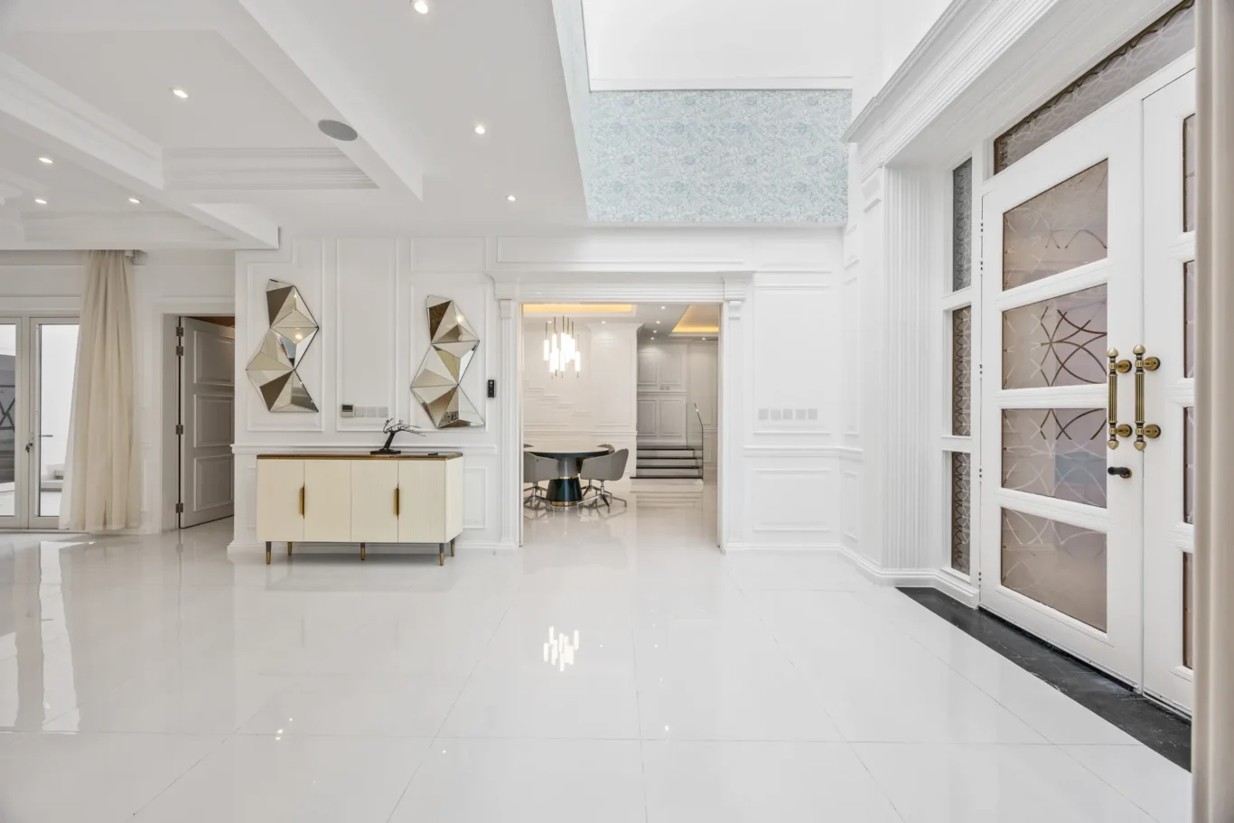 Bright white foyer with glossy tile floor, paneled walls, and geometric mirrored wall decor by a cream console.