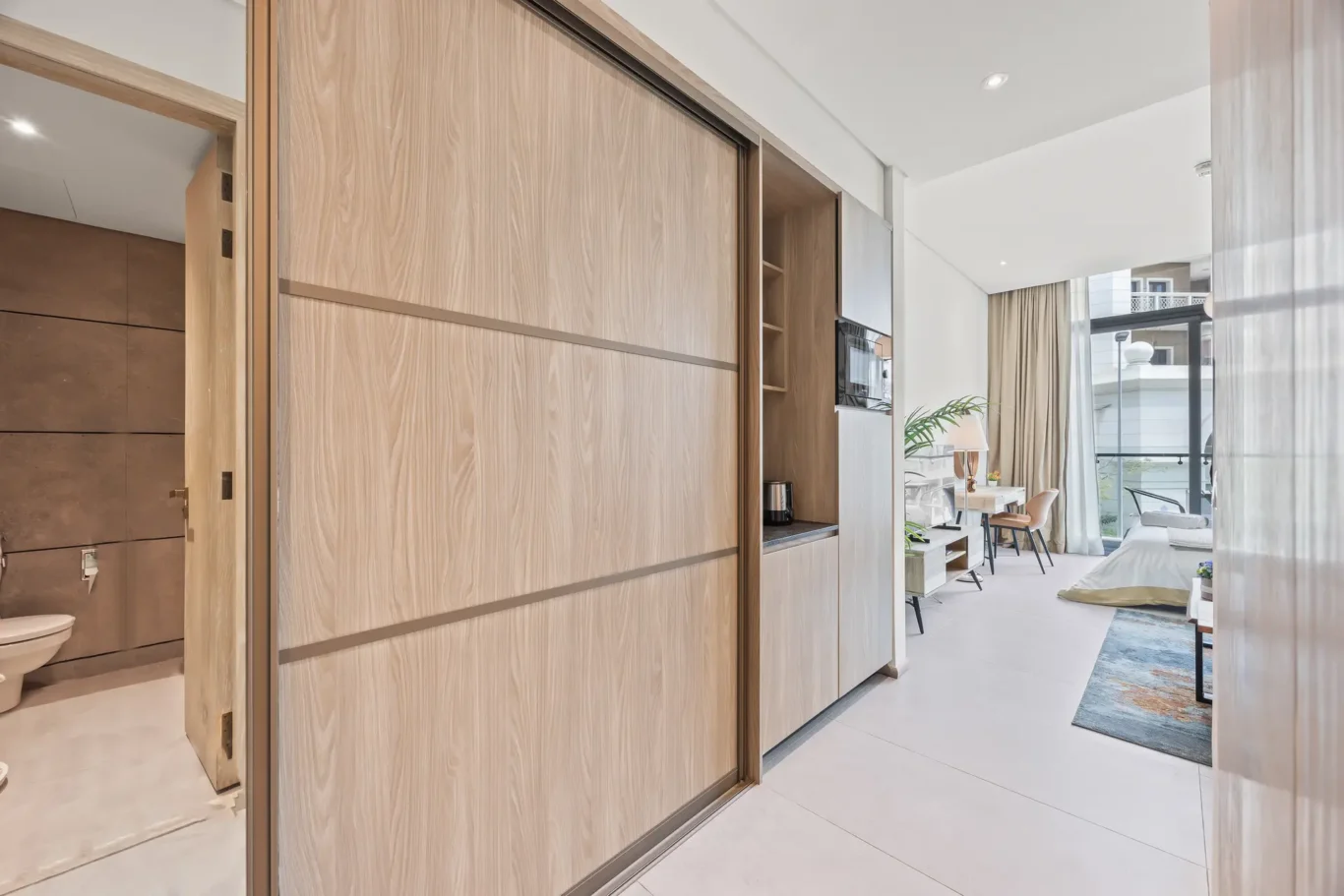 Modern hotel suite interior with tall wood sliding closets, open bathroom door on the left, and a bright living area with a desk and balcony view.