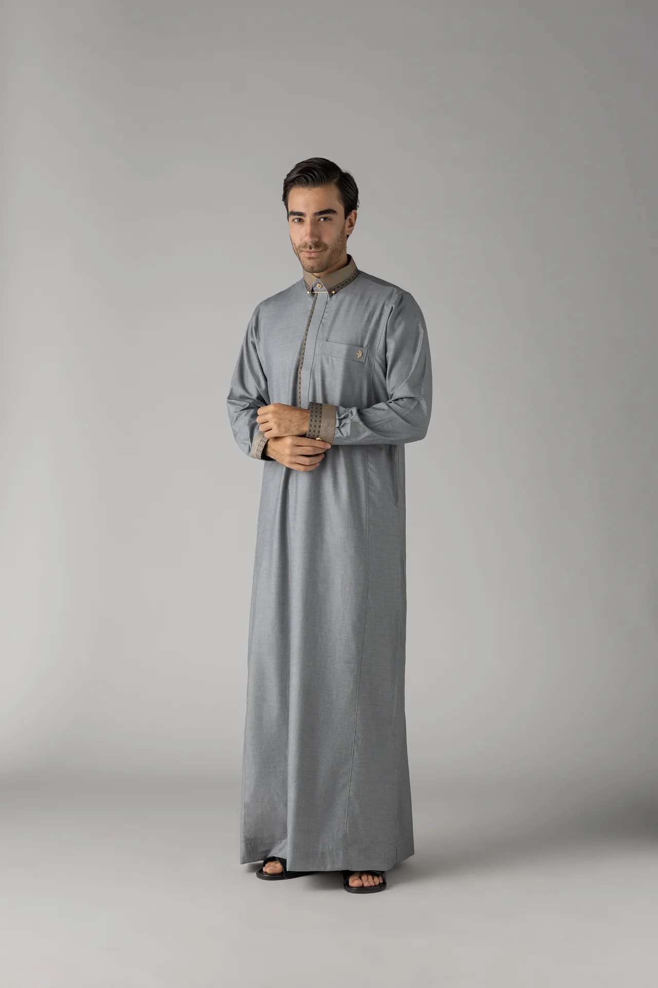Man standing in a light gray thobe, hands clasped, neutral studio backdrop.