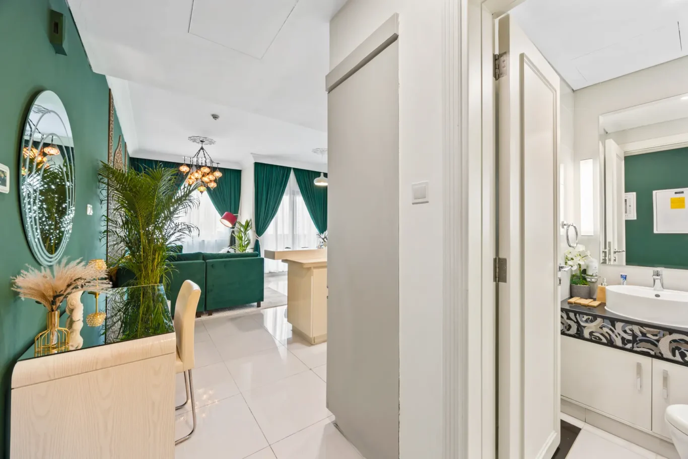 Open-concept living area with green walls, lush plants, and a chandelier over a teal sofa and cream furniture; doorway leads to a white-tiled bathroom.