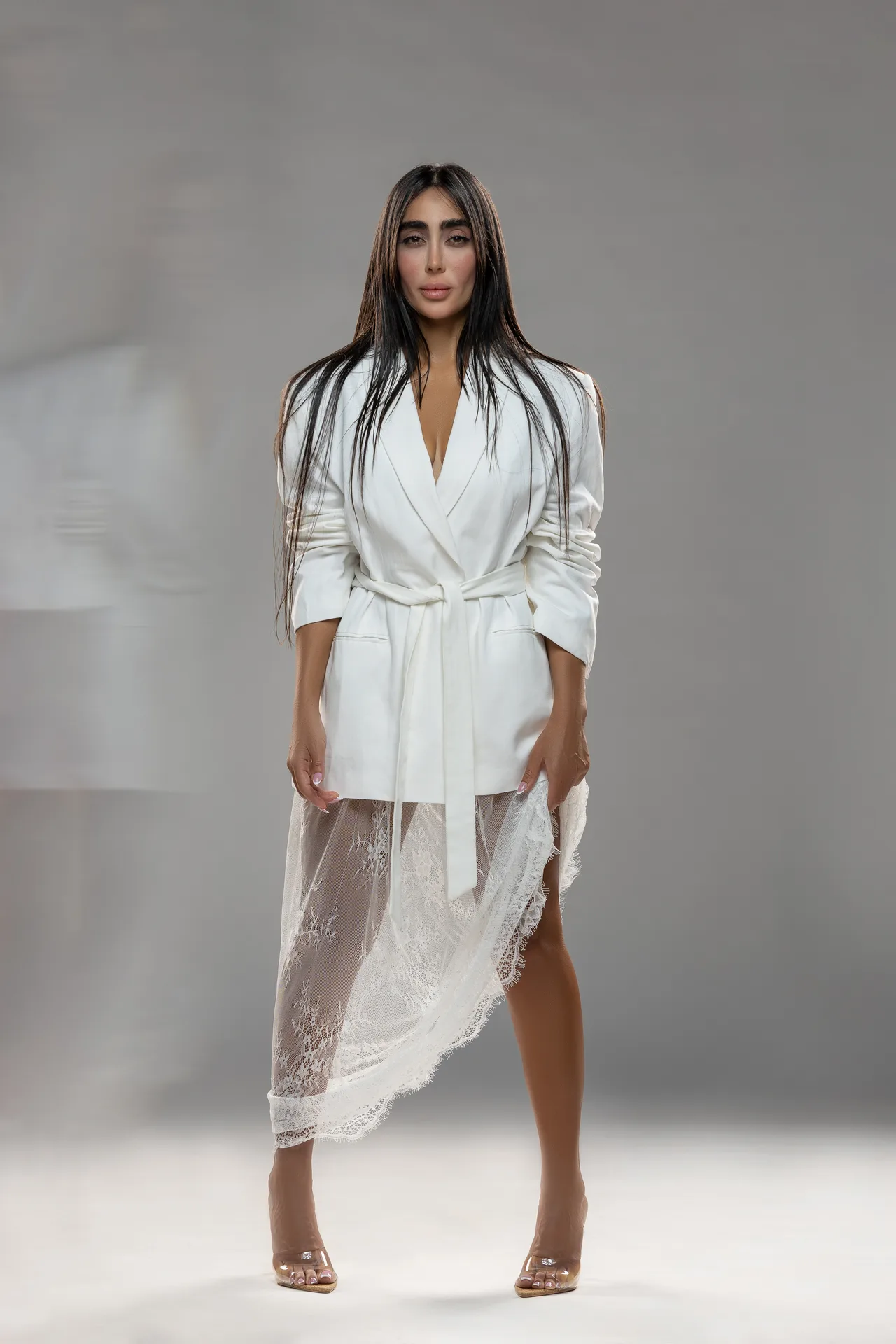 Runway model wearing a white belted robe with a sheer lace skirt and nude heels, posing against a gray studio background.