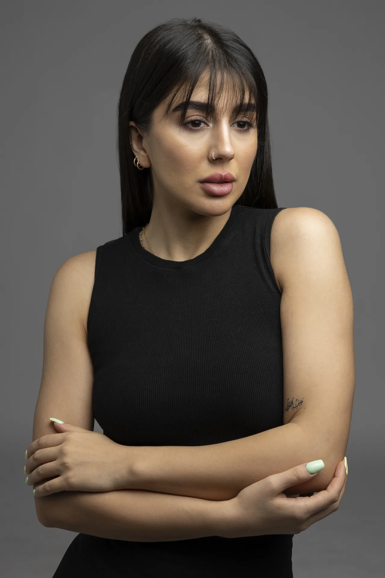 Woman with dark hair in a black sleeveless top, arms crossed, looking slightly to her right against a gray backdrop.