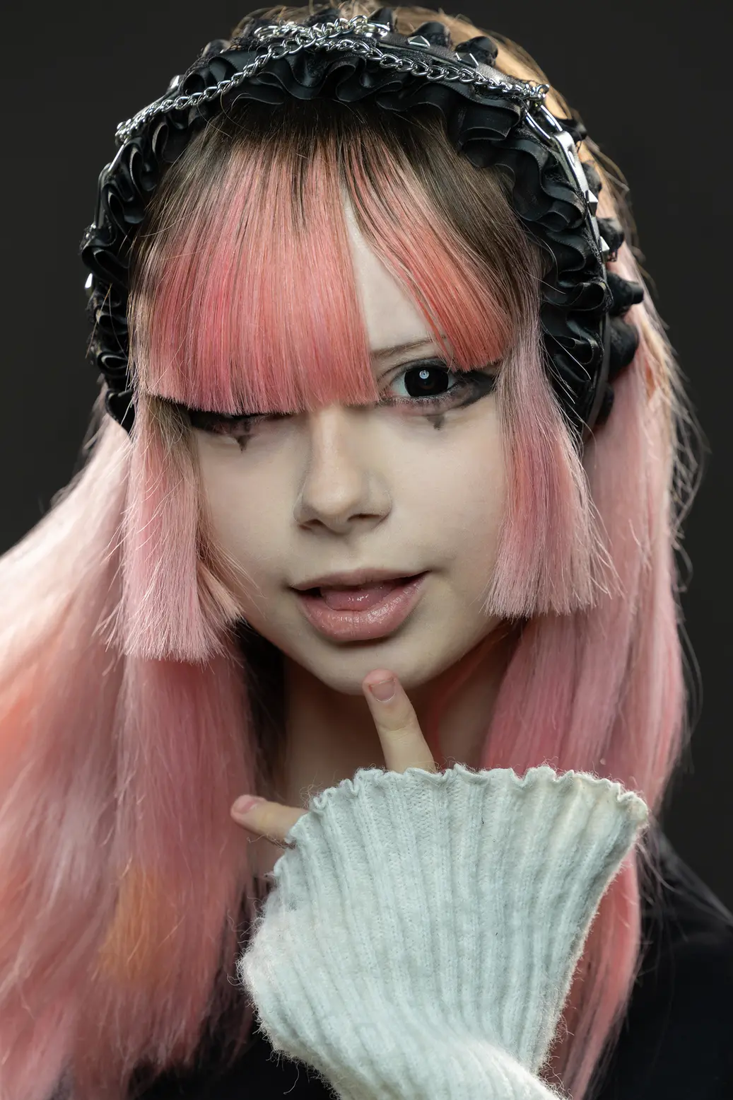 Portrait of a person with long pink hair and a black ruffled headband, winking and touching their lips with a pale sweater sleeve in the foreground.