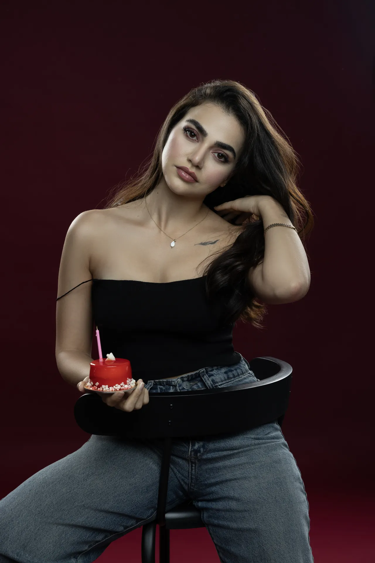Woman sitting on a black chair holding a red cupcake with a candle against a dark burgundy background.