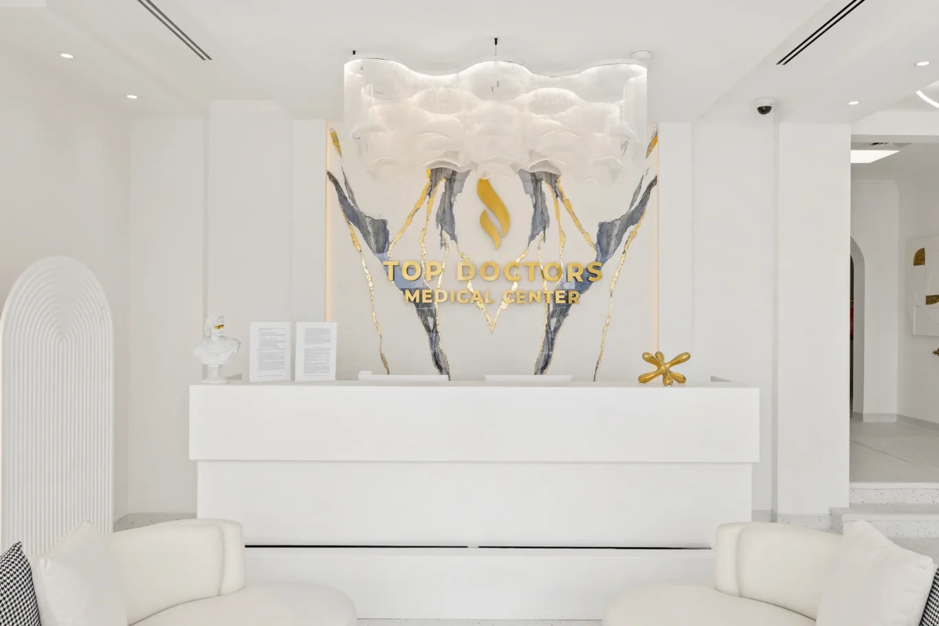 White modern medical reception area with a marble wall, gold-accent logo reading 'Top Doctors Medical Center', and a white reception desk behind it.