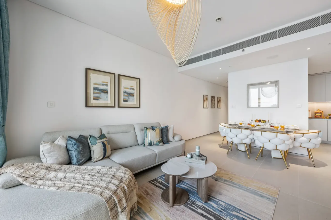 Bright open-plan living and dining area: light gray sectional sofa with assorted pillows, two round coffee tables, and a dining table with white tufted chairs and gold legs set for a meal.