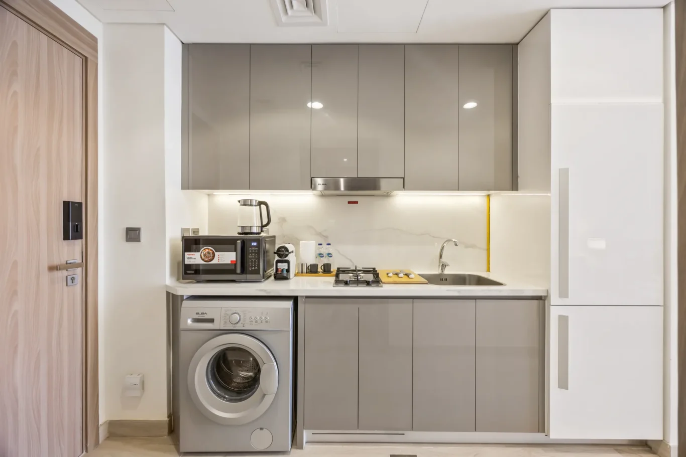 Compact modern kitchen with grey glossy upper cabinets, white countertop, and built-in appliances (microwave, kettle, sink, stove).