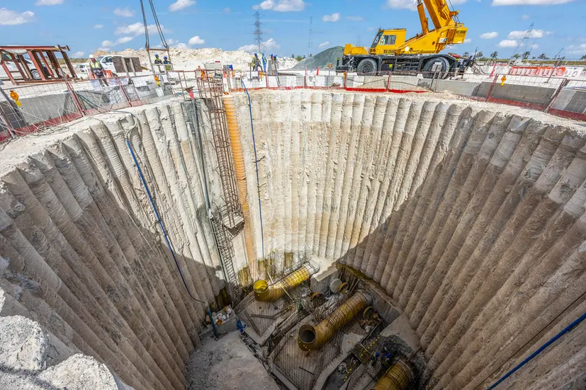 Large circular excavation pit with reinforced concrete walls and yellow drainage pipes; construction site with a crane and workers in the background.