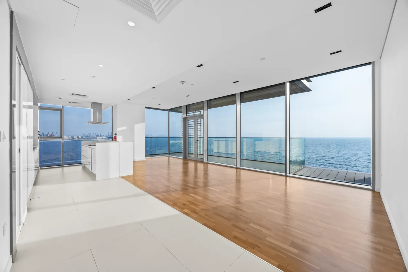 Open-concept living area with floor-to-ceiling ocean-view windows, white walls, and a wooden floor. A kitchen island is on the left.
