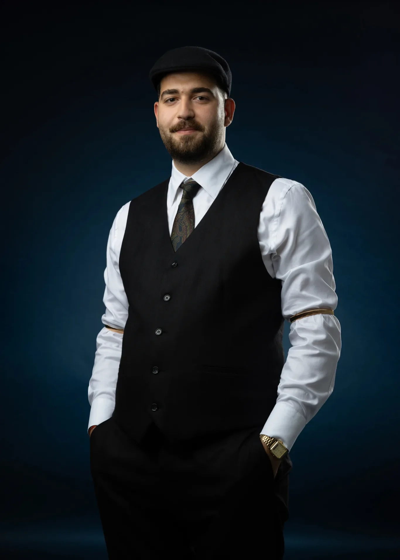 Portrait of a man in a black waistcoat, white shirt, and paisley tie, wearing a flat cap, against a dark blue gradient background, looking at the camera.