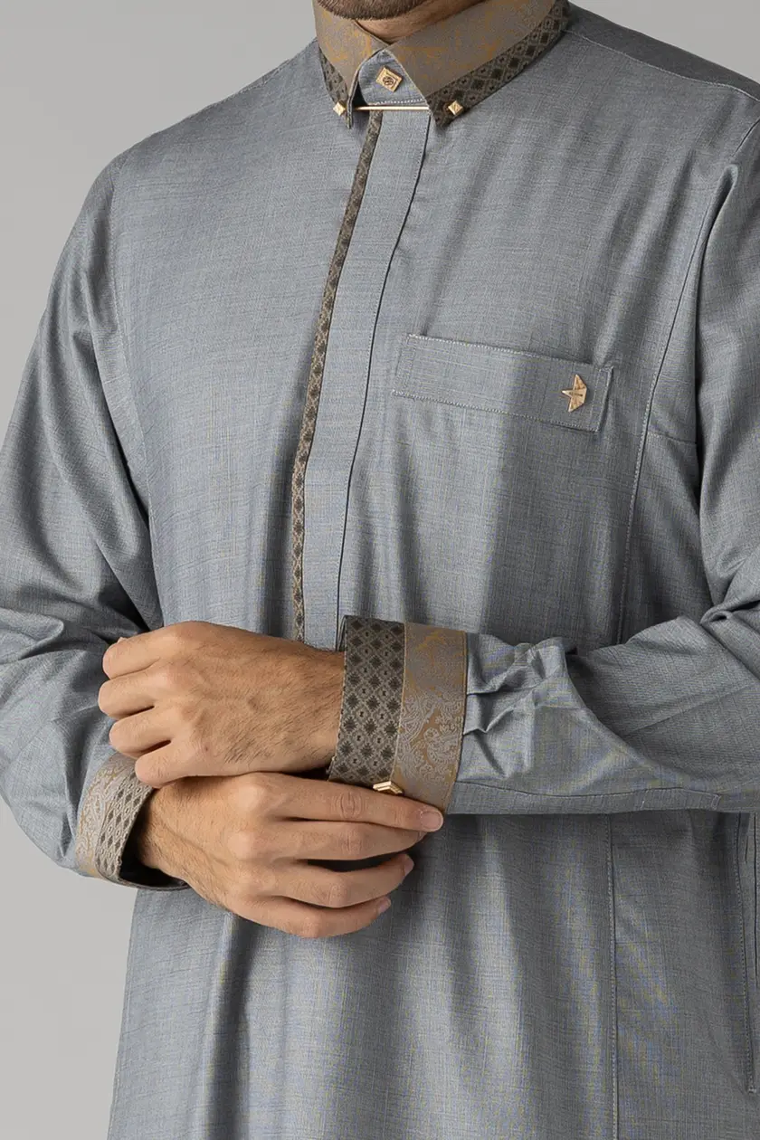 Close-up of a man wearing a light gray thobe with brown patterned collar and cuffs, adjusting his sleeve.