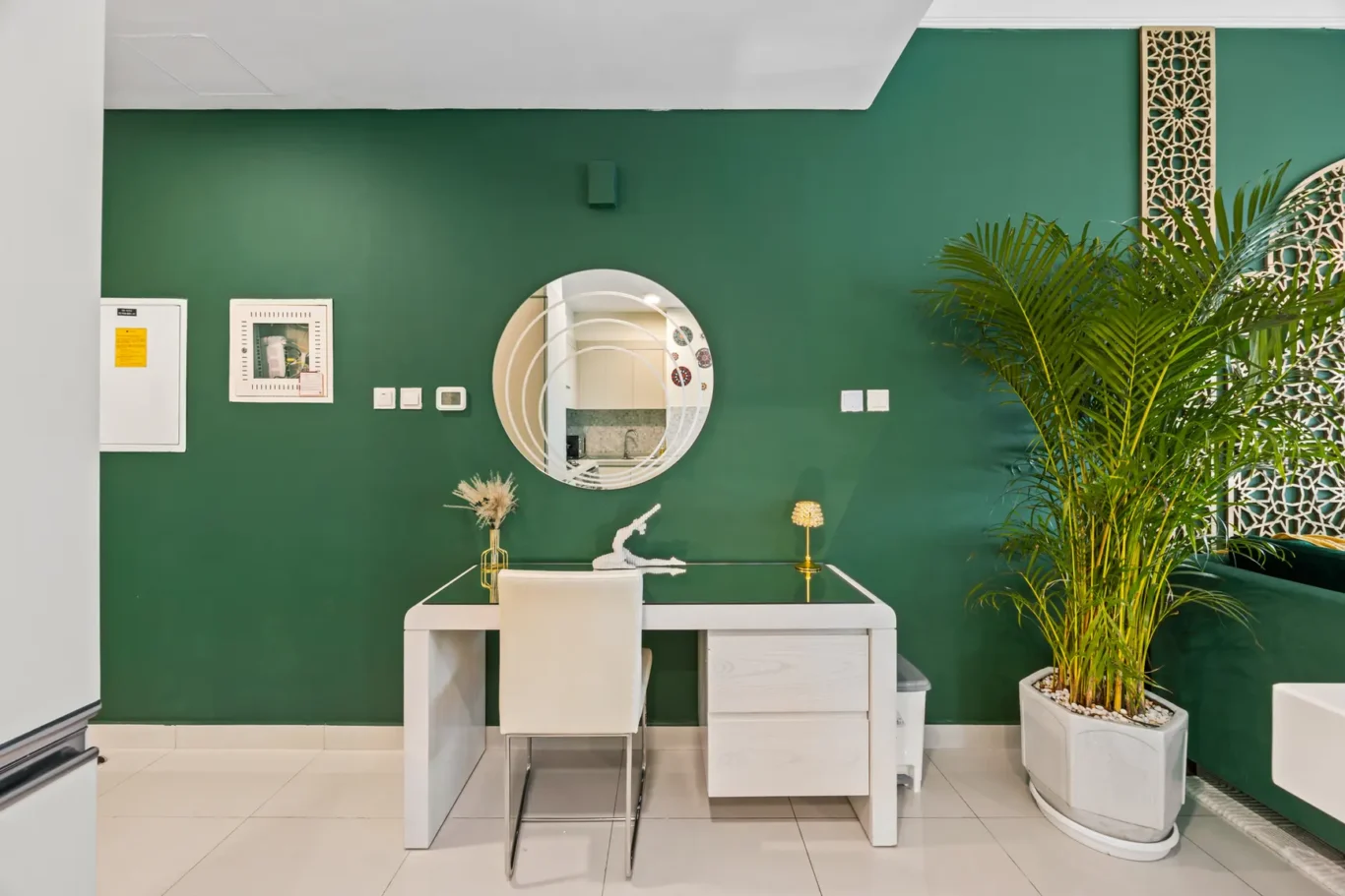Modern white desk against a dark green wall with a round mirror, decorative items, and a large potted plant nearby.