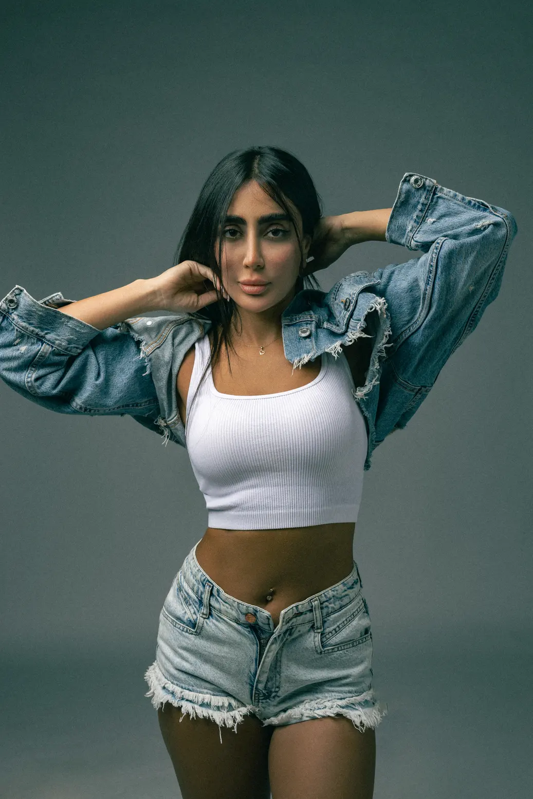 Woman posing in a casual denim outfit: white crop top, distressed denim shorts, denim jacket draped over shoulders, hands in hair against a neutral studio background.