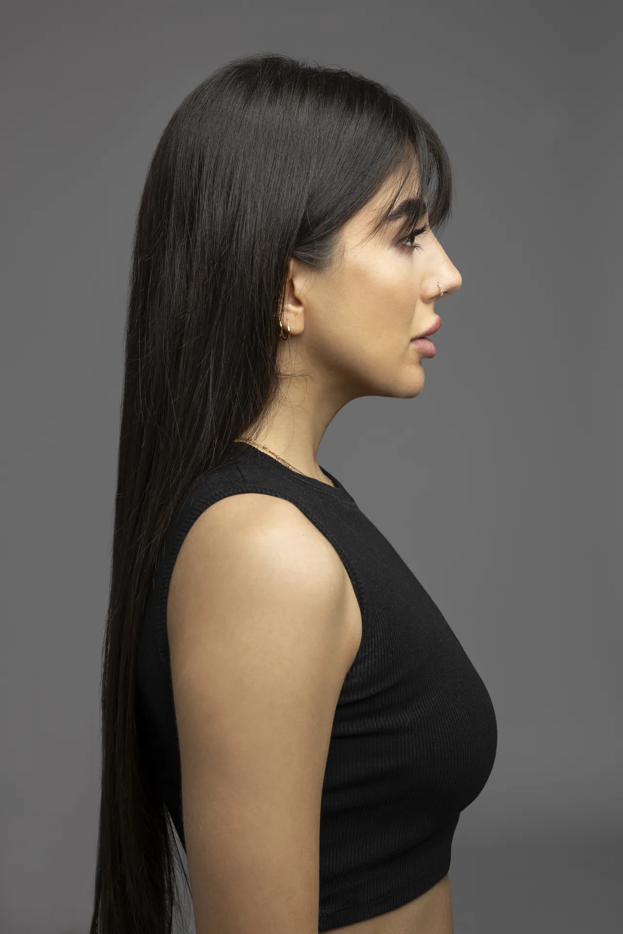 Profile view of a woman with long straight black hair, wearing a black sleeveless top against a gray studio background, looking to the right.