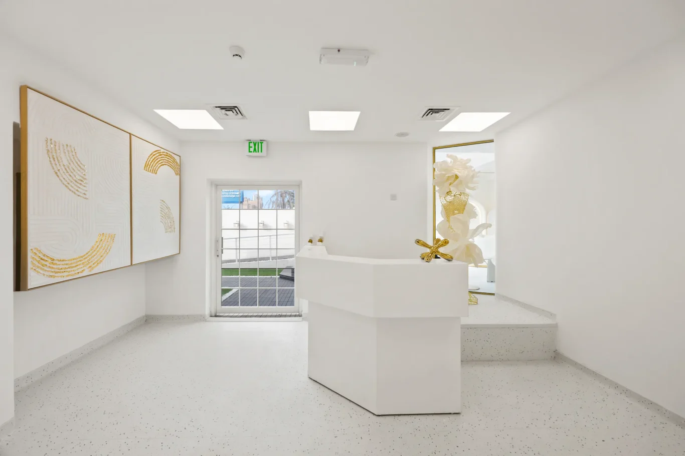 White modern lobby with abstract gold-art panels on the left and a white reception desk in the center; glass exit door ahead and decorative gold sculpture near the door