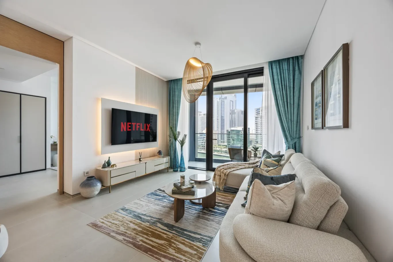 Living room with a wall-mounted Netflix screen above a white console, beige sofa, and a colorful rug facing a glass balcony with city buildings outside.",