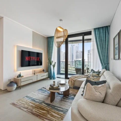 Living room with a wall-mounted Netflix screen above a white console, beige sofa, and a colorful rug facing a glass balcony with city buildings outside.",