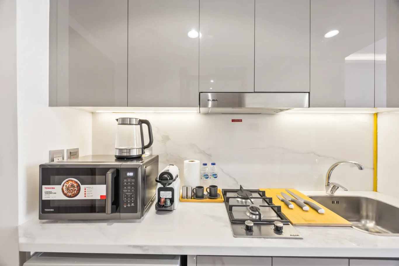 Modern kitchen counter with microwave, electric kettle, coffee machine, and a knife board beside a gas stove near a sink.