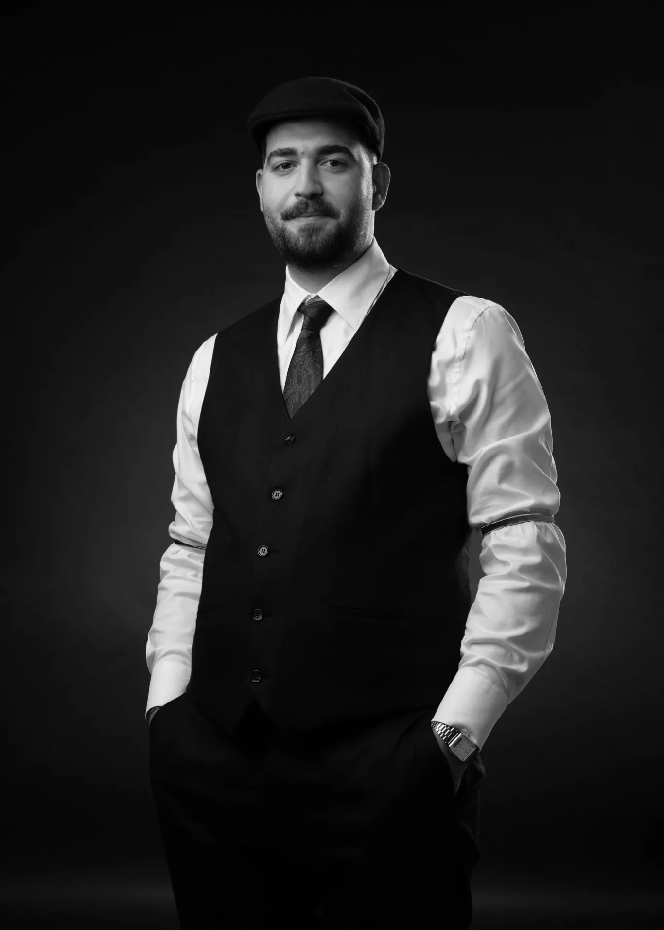 Black-and-white portrait of a man in a dark waistcoat, white shirt, patterned tie, and flat cap, standing against a dark studio backdrop with hands in pockets.