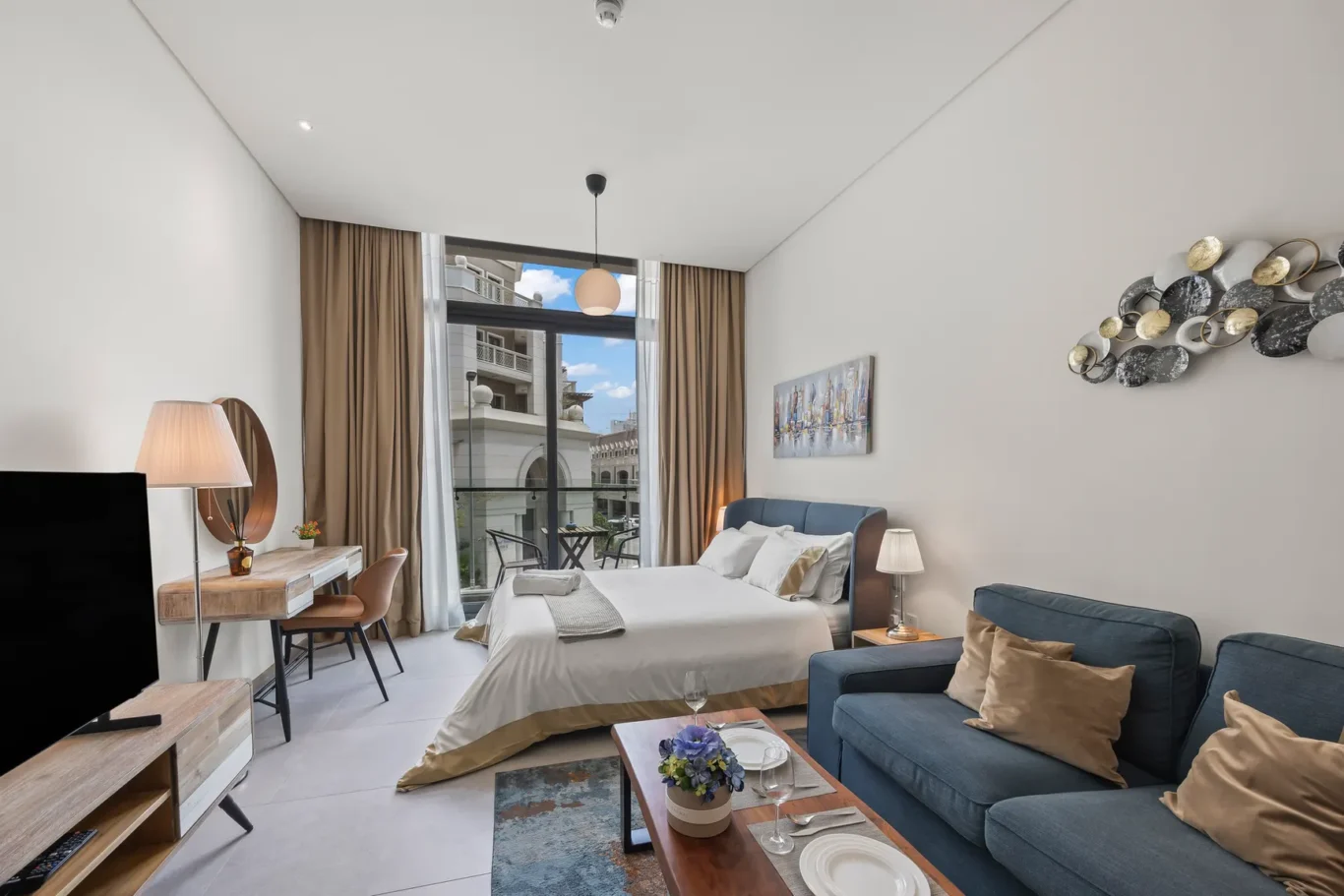 Bright hotel suite with a king bed, blue upholstered headboard, and a seating area by a large window balcony.