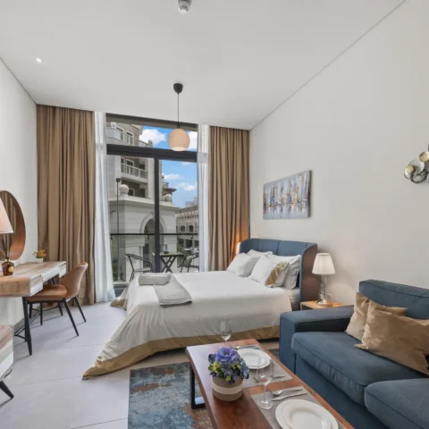 Bright hotel suite with a king bed, blue upholstered headboard, and a seating area by a large window balcony.