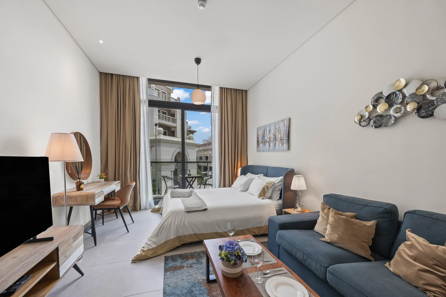Bright hotel suite with a king bed, blue upholstered headboard, and a seating area by a large window balcony.
