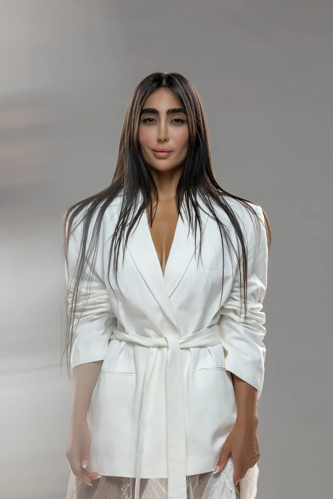 Woman wearing a white belted blazer over a lace skirt, standing against a gray studio backdrop with a neutral expression.