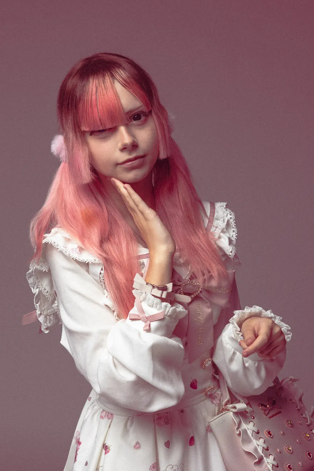 Person with long pink hair in a pastel Lolita-style white dress, posing with hand under the chin against a pink backdrop.