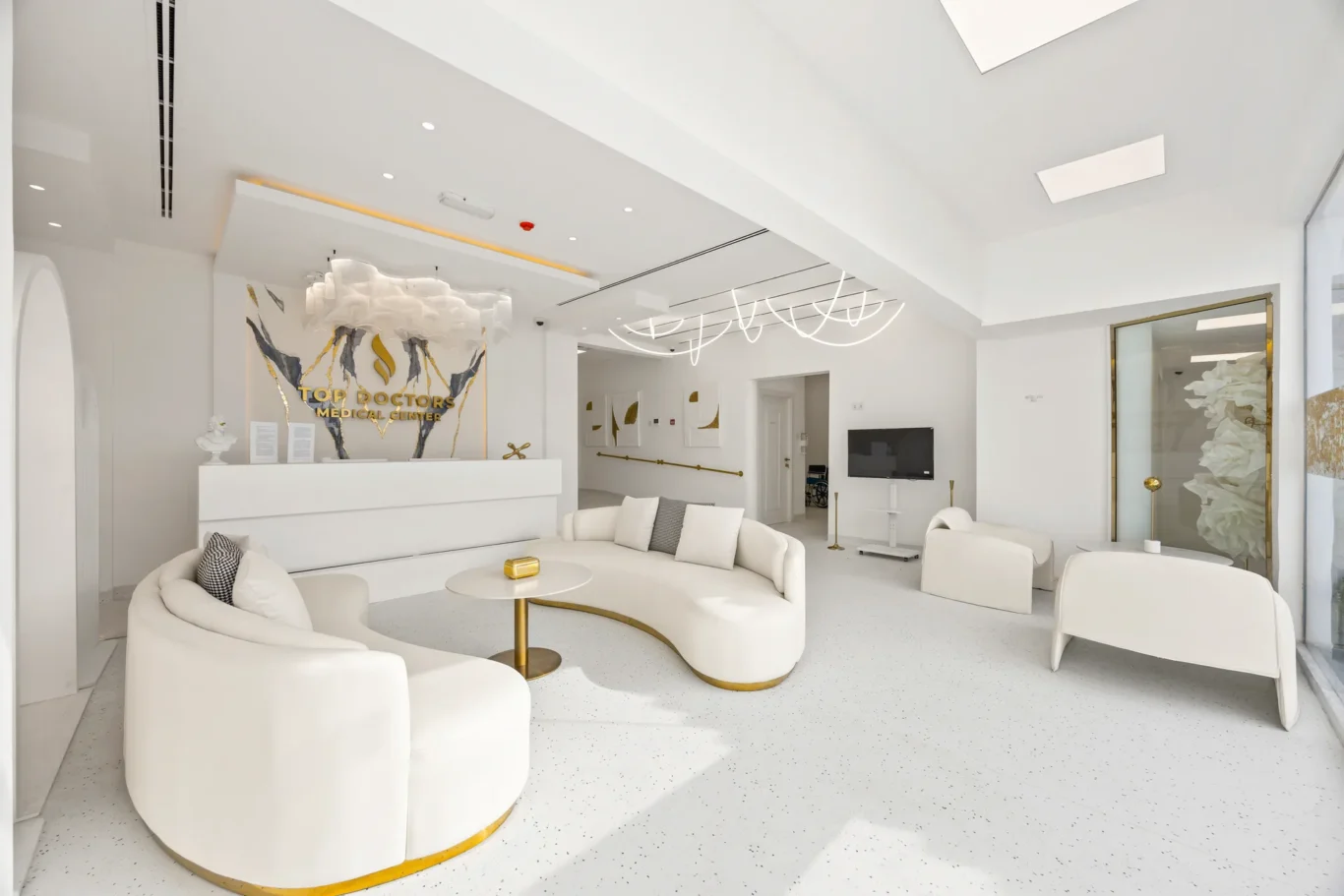 Bright white medical lobby with curved white seating and gold accents, reception desk, and modern sculptures.