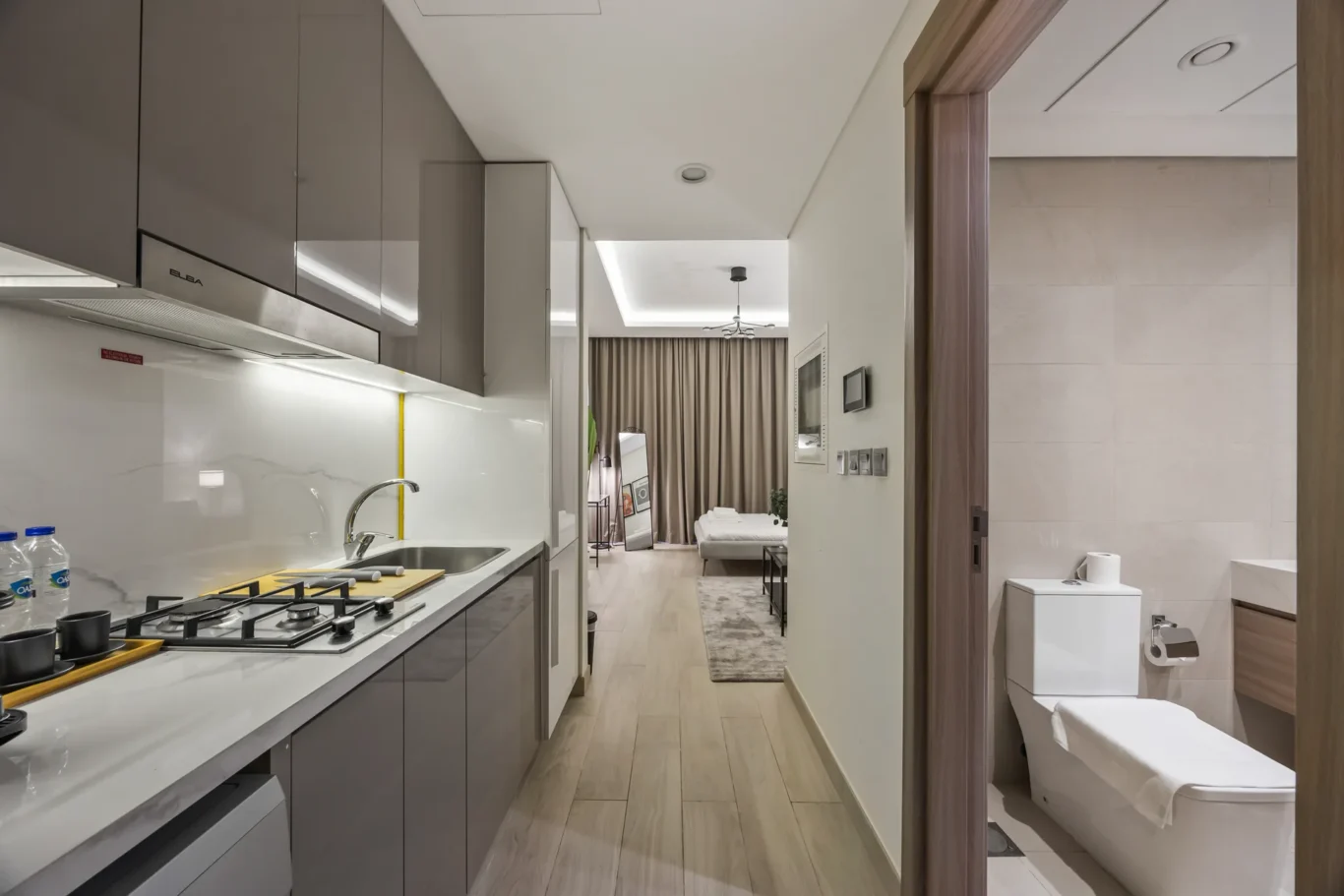 Open-plan kitchen with grey cabinets and gas stove leading to a living area with a bed and brown curtains in the background.