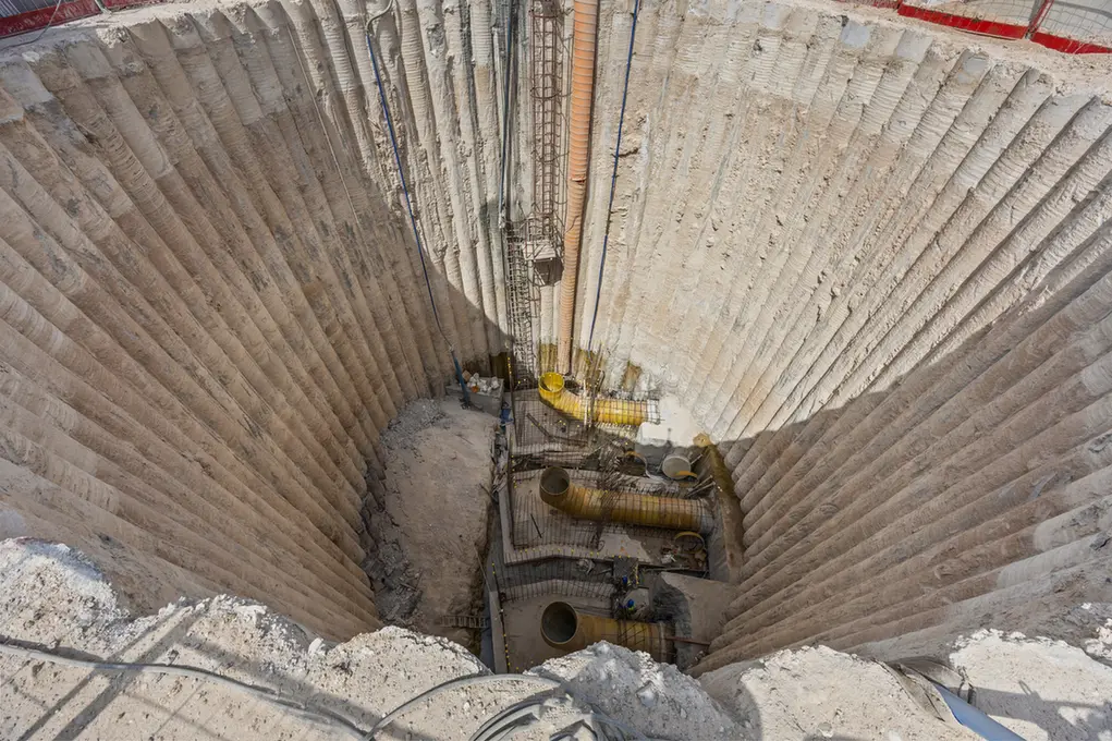 Deep circular construction shaft with reinforced concrete walls, exposed pipes, and equipment at the bottom.