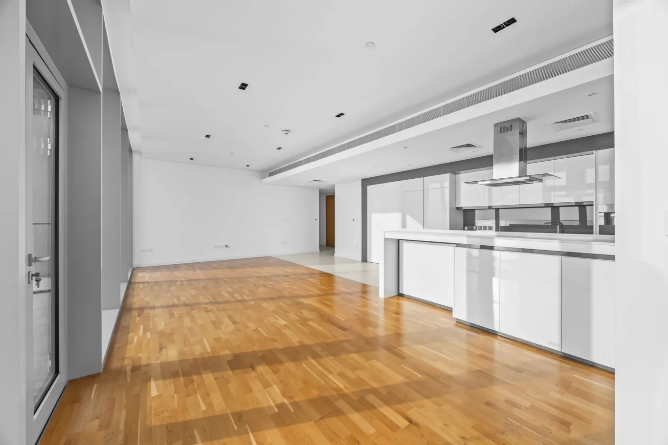 Open-plan kitchen and living area with white cabinets, stainless steel range hood, and a wooden parquet floor; bright, empty space.