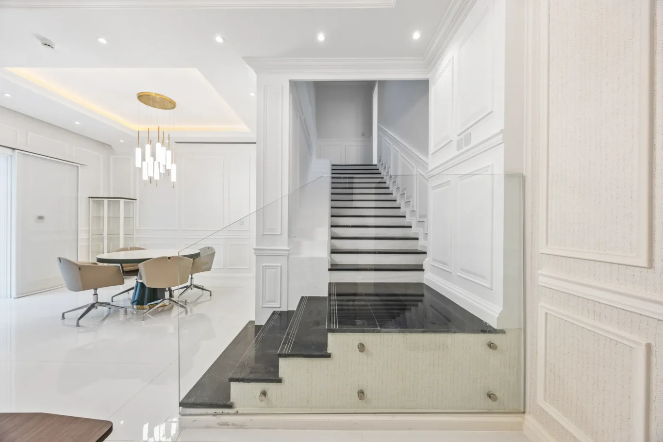 Bright white interior with a black marble staircase and glass railing, a round dining table and beige chairs to the left, and a gold pendant chandelier above.