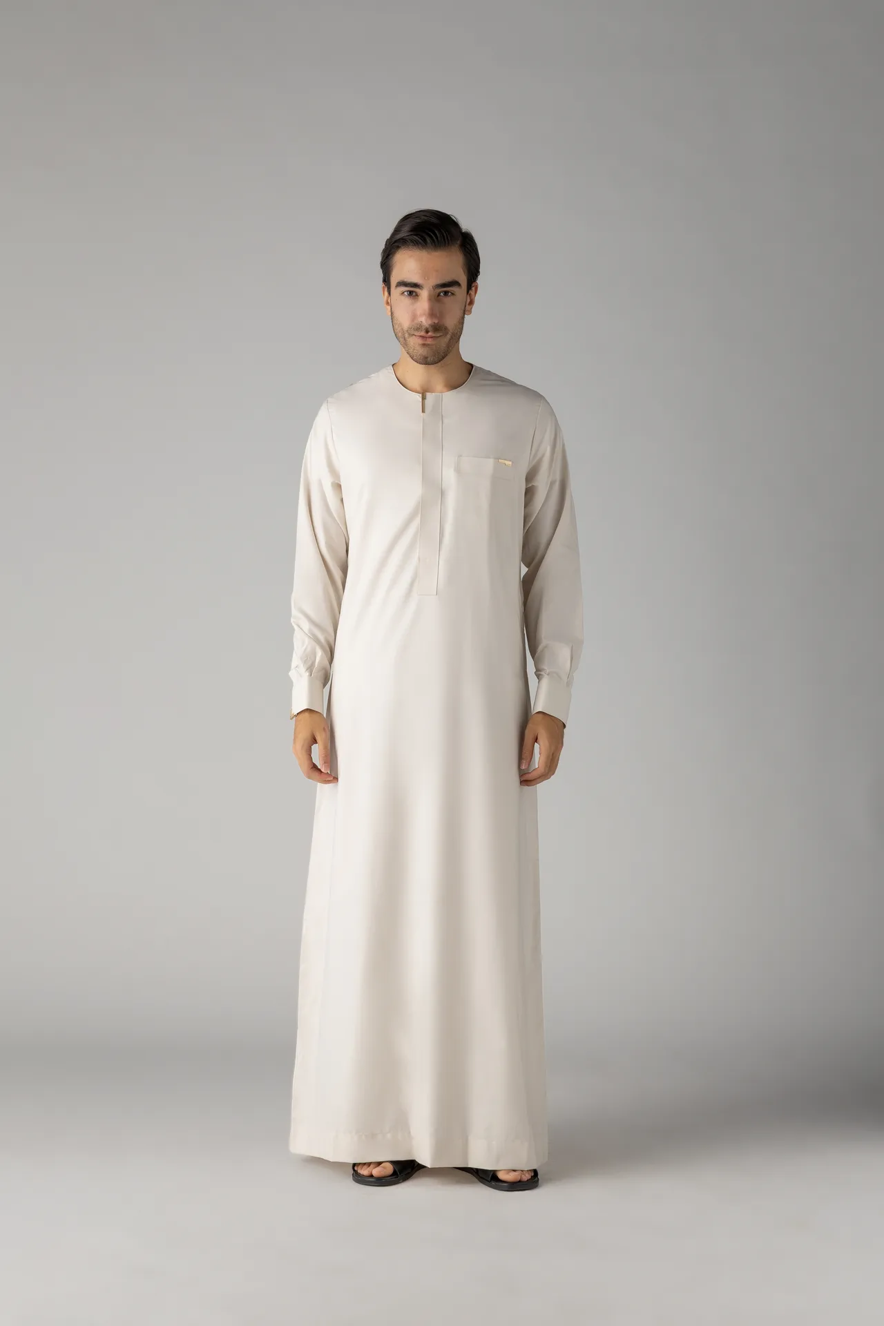 Man wearing a cream-colored thobe with a chest pocket, standing against a gray backdrop.