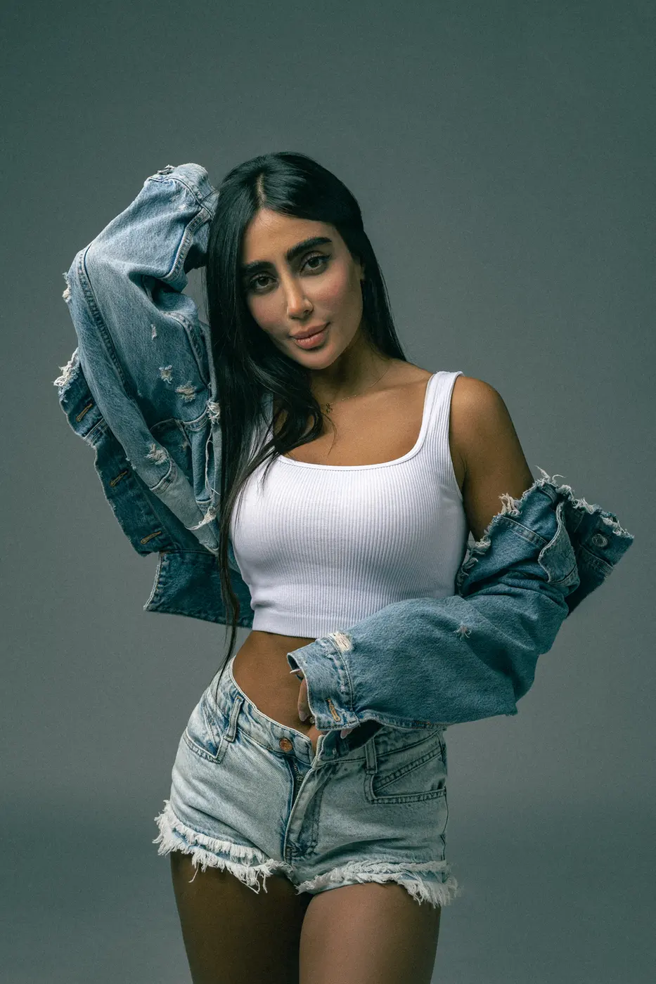 Woman in a white ribbed tank top and frayed denim shorts, with a distressed denim jacket draped over her shoulders, posing against a gray backdrop.