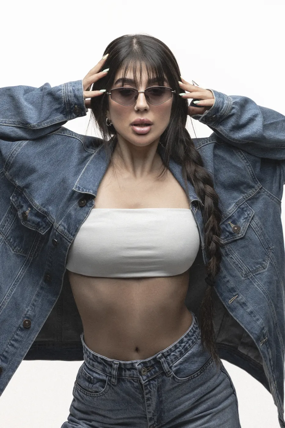 Woman wearing a denim jacket and jeans with a white tube top, sunglasses, and a long braided ponytail posing with hands in her hair against a white studio background.