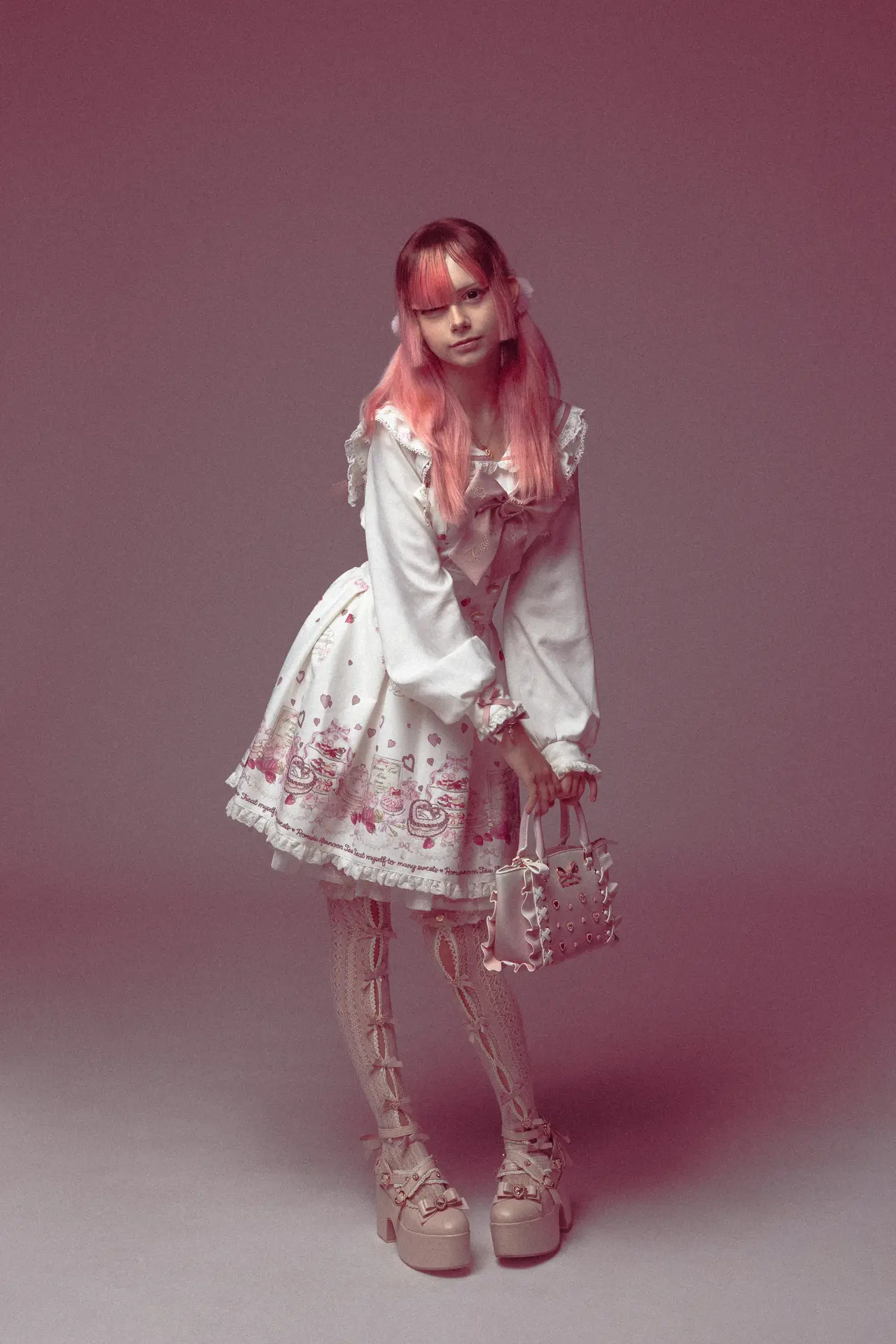 Person in pastel Lolita dress with pink wig, holding a pink handbag, standing against a pink backdrop.