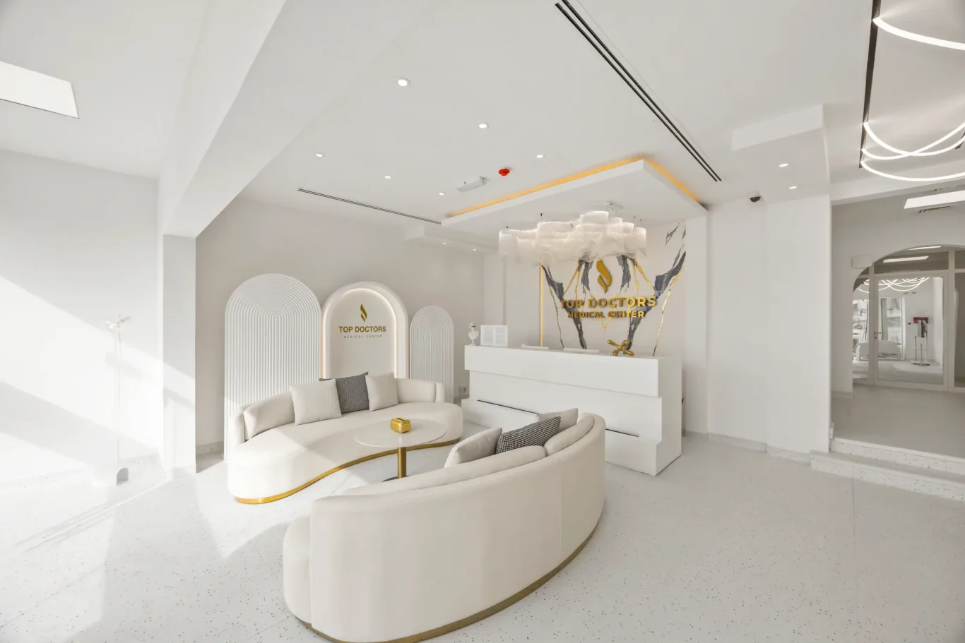 Bright white medical reception area with curved white seating, gold-trimmed coffee table, and a reception desk featuring 'Top Doctors Medical Center' signage in gold on a decorative backdrop.