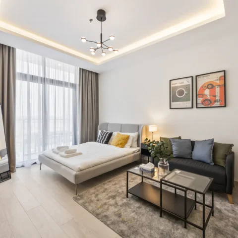 Modern hotel-style bedroom with gray upholstered bed, a sofa, and a glass coffee table on a gray rug; large windows with sheer and blackout curtains on the left; green plant in foreground and two framed art prints above the bed.