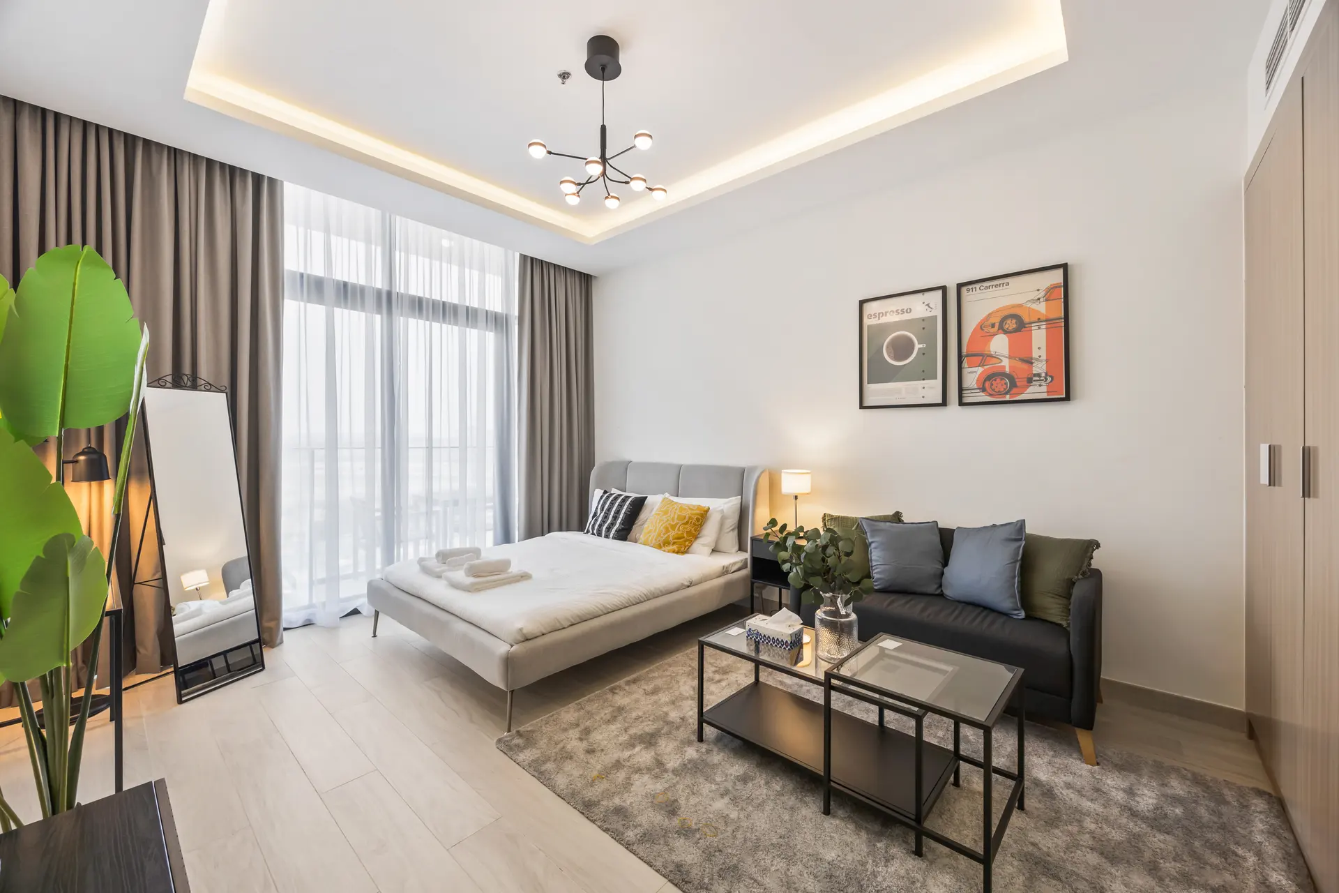 Modern hotel-style bedroom with gray upholstered bed, a sofa, and a glass coffee table on a gray rug; large windows with sheer and blackout curtains on the left; green plant in foreground and two framed art prints above the bed.