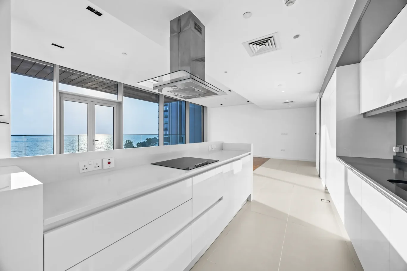 Bright modern white kitchen with a long island, built-in stove, and a view of the ocean through large glass doors and windows.