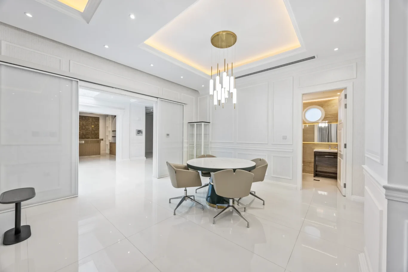 White dining room with a round glass table, beige chairs, and a gold pendant chandelier in a glossy white space.