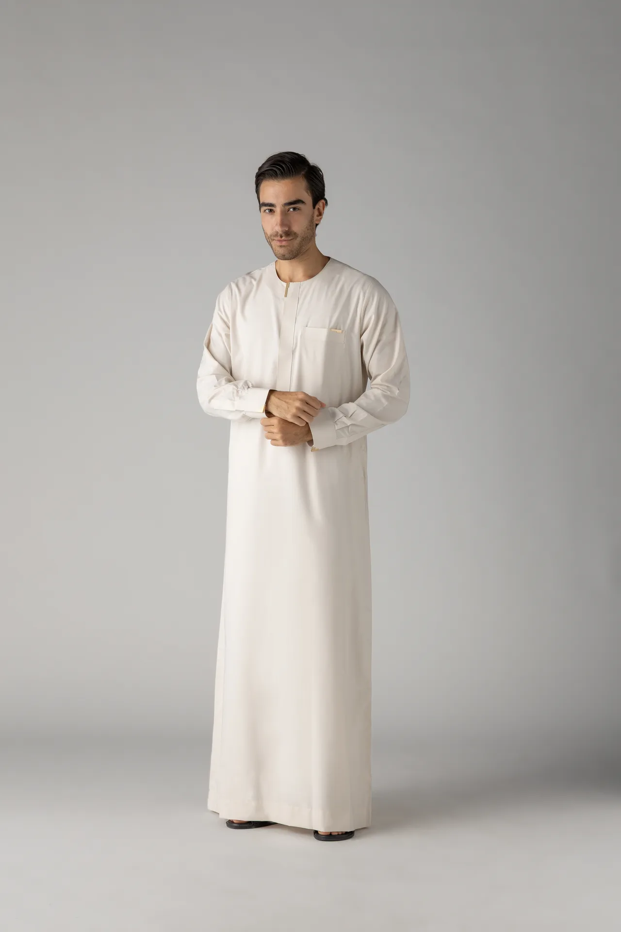 Man wearing a white thobe/robe stands against a neutral gray studio backdrop with hands clasped in front.
