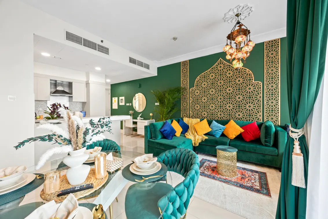 Open-concept living and dining area with emerald-green velvet sofa, colorful throw pillows, and a gold geometric wall panel backdrop."Note: elegant chandelier above" (informative image)