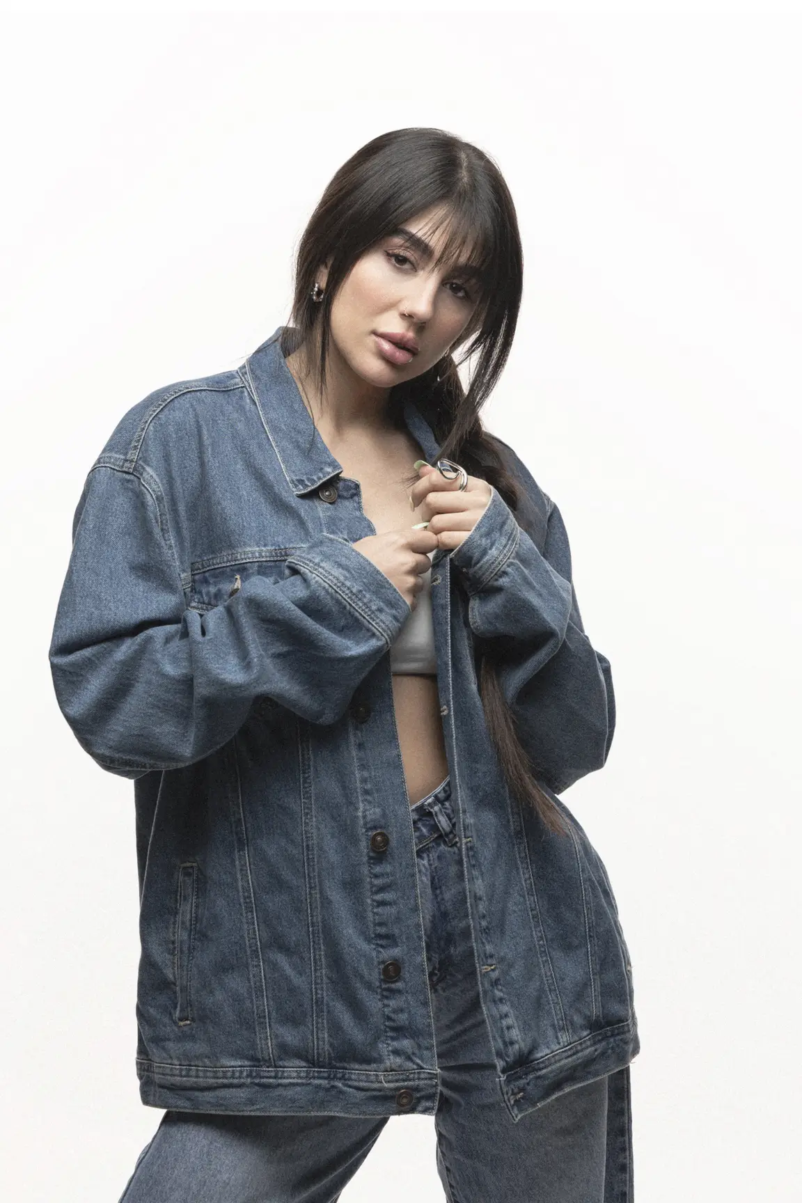 Young woman with long dark hair wearing an oversized denim jacket over a white top, posing against a light background.