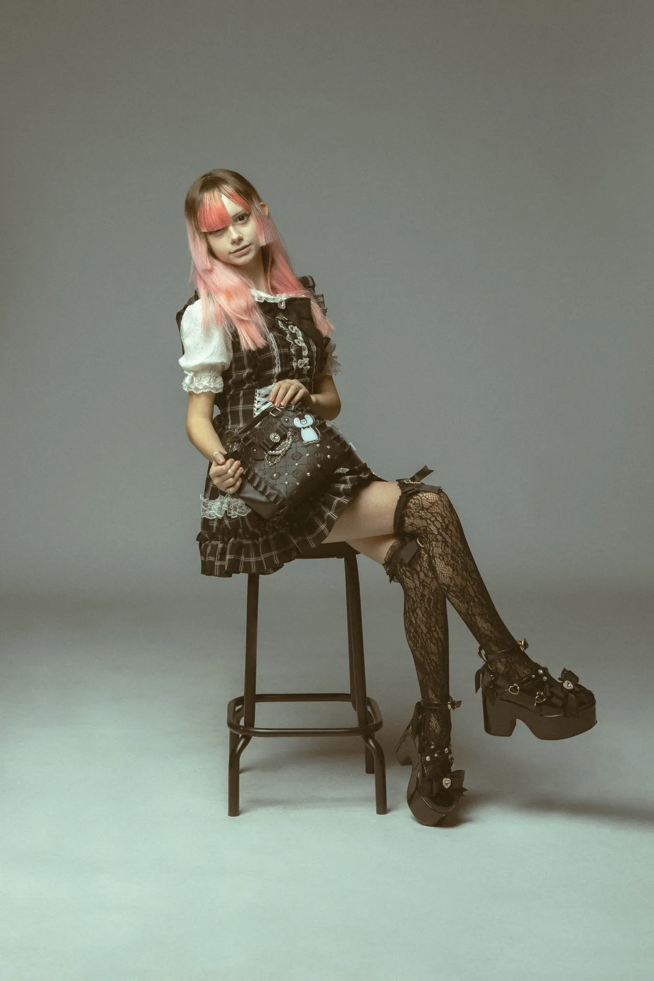 Pink-haired young woman in a plaid lacy dress sits on a stool, holding a black studded handbag in a studio setting.