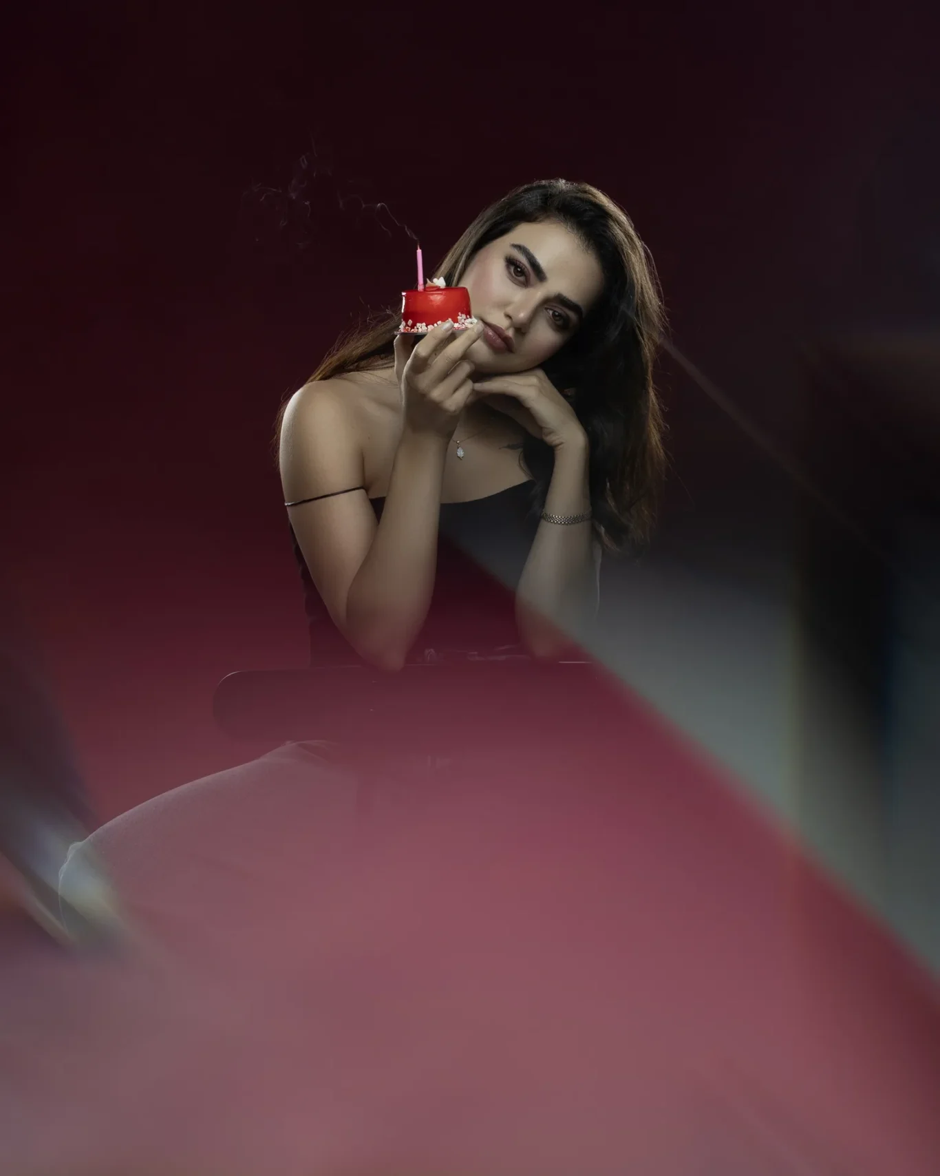 Woman in a black off-shoulder top leans on her hand while holding a small red cake with a pink candle close to her face, against a dark background.