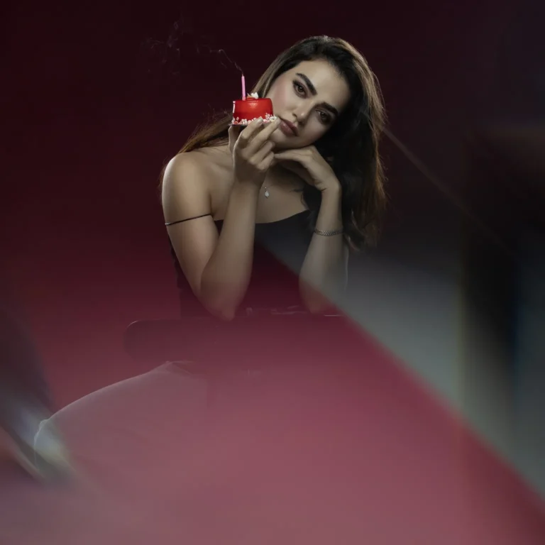 Woman in a black off-shoulder top leans on her hand while holding a small red cake with a pink candle close to her face, against a dark background.