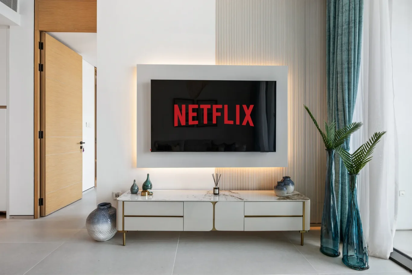 Modern living room with a wall-mounted TV showing the Netflix logo, centered above a white marble-top cabinet with gold trim. Tall teal vases and sheer curtains to the right amplify the stylish space.